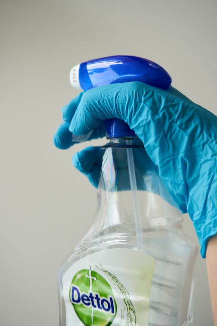 A close-up of a hand wearing a bright blue disposable glove, holding a transparent spray bottle filled with a clear cleaning solution. The spray nozzle, which is blue and white, is being pressed to di